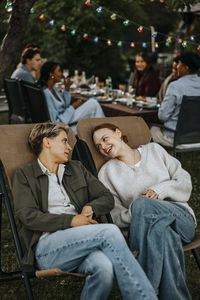 Female friends with legs crossed at knee sitting on chair and talking in back yard at social party