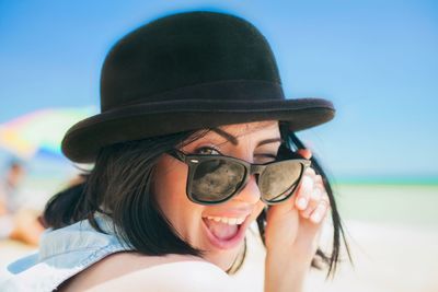 Portrait of cheerful woman wearing sunglasses winking at beach against sky