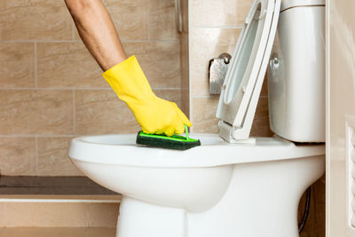 Human hand in a yellow rubber glove is using a plastic brush to scrub the toilet bowl.