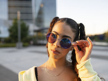 Confident young fit ethnic female wearing sunglasses while relaxing on the street in city park and looking away