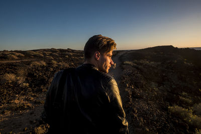 Rear view of thoughtful young man looking away while standing on land against sky during sunset