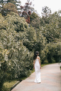 An elegant young woman bride in a wedding dress walks through a green park among plants and trees