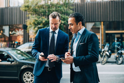 Mature male business colleagues using mobile phones while standing on sidewalk in city