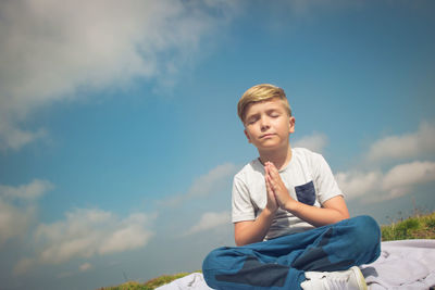 Small boy in namaste position meditating with eyes closed on a meadow against the sky. copy space.