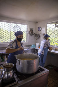 Serious adult ethnic female cook adding herbs in big saucepan while cooking in restaurant kitchen in colombia