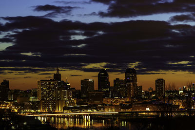 Illuminated buildings by river against sky at sunset