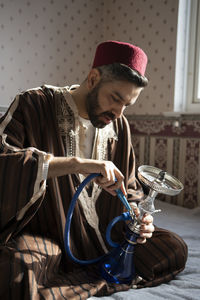 Man in traditional wear holding hookah at home