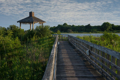 Wooden boardwalk leading towards gazebo amidst plants against sky on sunny day