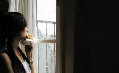 Thoughtful woman with flower looking through window during rainy season