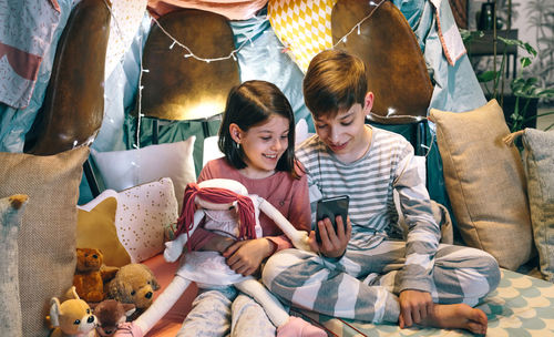 Happy children laughing together while watching video on cell phone sitting on shelter tent at home