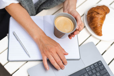 A woman works on a laptop, takes notes, and drinks coffee during a meeting in a cafe.