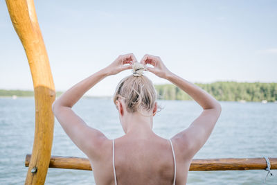 Rear view of woman tying hair by lake against sky