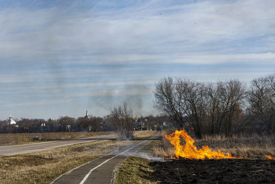 Road by bare trees on field against sky with fire