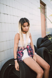 Young woman looking down while sitting on tire against tiled wall