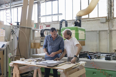 Project manager and coworker working while standing against sawing machine at factory