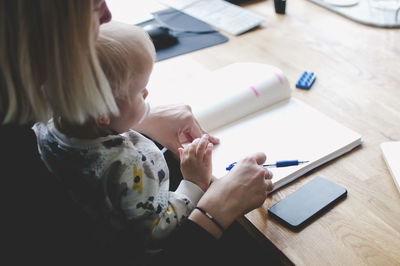 High angle view of businesswoman with baby girl writing in diary at table in creative office