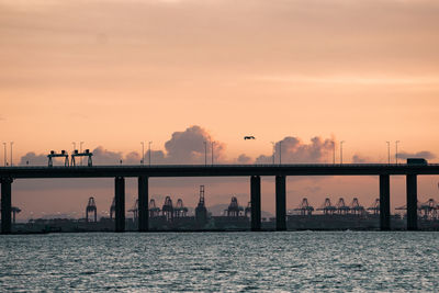 Silhouette factory by sea against sky during sunset