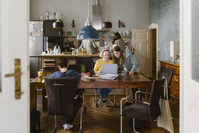 Grandmother using laptop with granddaughter at dining table in home seen through doorway