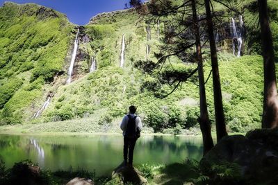 Rear view of man standing at lakeshore against waterfall