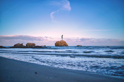Woman standing on rock in sea against sky during sunset