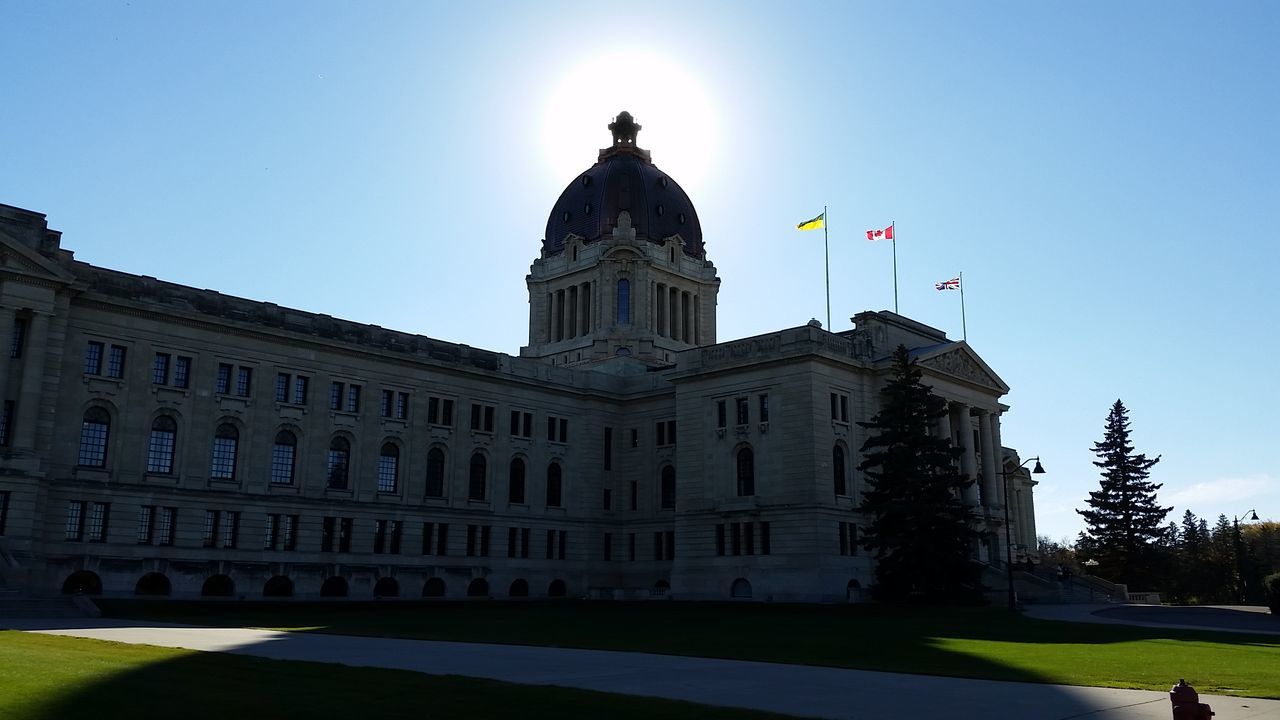 architecture, building exterior, built structure, clear sky, flag, sunlight, low angle view, dome, blue, facade, city, outdoors, travel destinations, day, sun, lawn, arch, city life, footpath, government building, sky, history, architectural feature, majestic, parliament building, arcade, spire