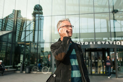 Senior male commuter looking away while talking through smart phone against railroad station building