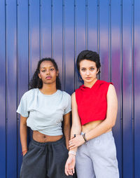 Serious multiracial young female friends in casual clothes looking at camera while standing together against blue background on street of city