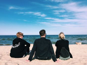 Rear view of young friends sitting at beach against sky