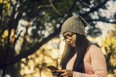 Low angle view of woman using mobile phone while standing against tree