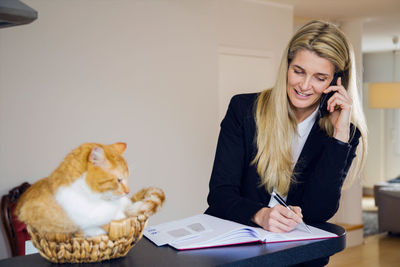 Mature businesswoman using mobile phone while writing in book by cat sitting in basket at home