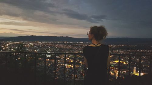 Rear view of woman looking at illuminated cityscape against sky during sunset