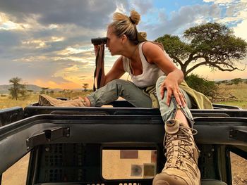 Full length of woman looking through binoculars while sitting on car against sky