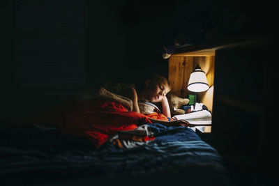 Boy reading book in darkroom under electric lamp at home