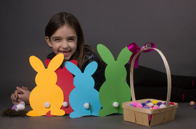 Portrait of smiling girl with eater bunny and pom pom in basket against gray background