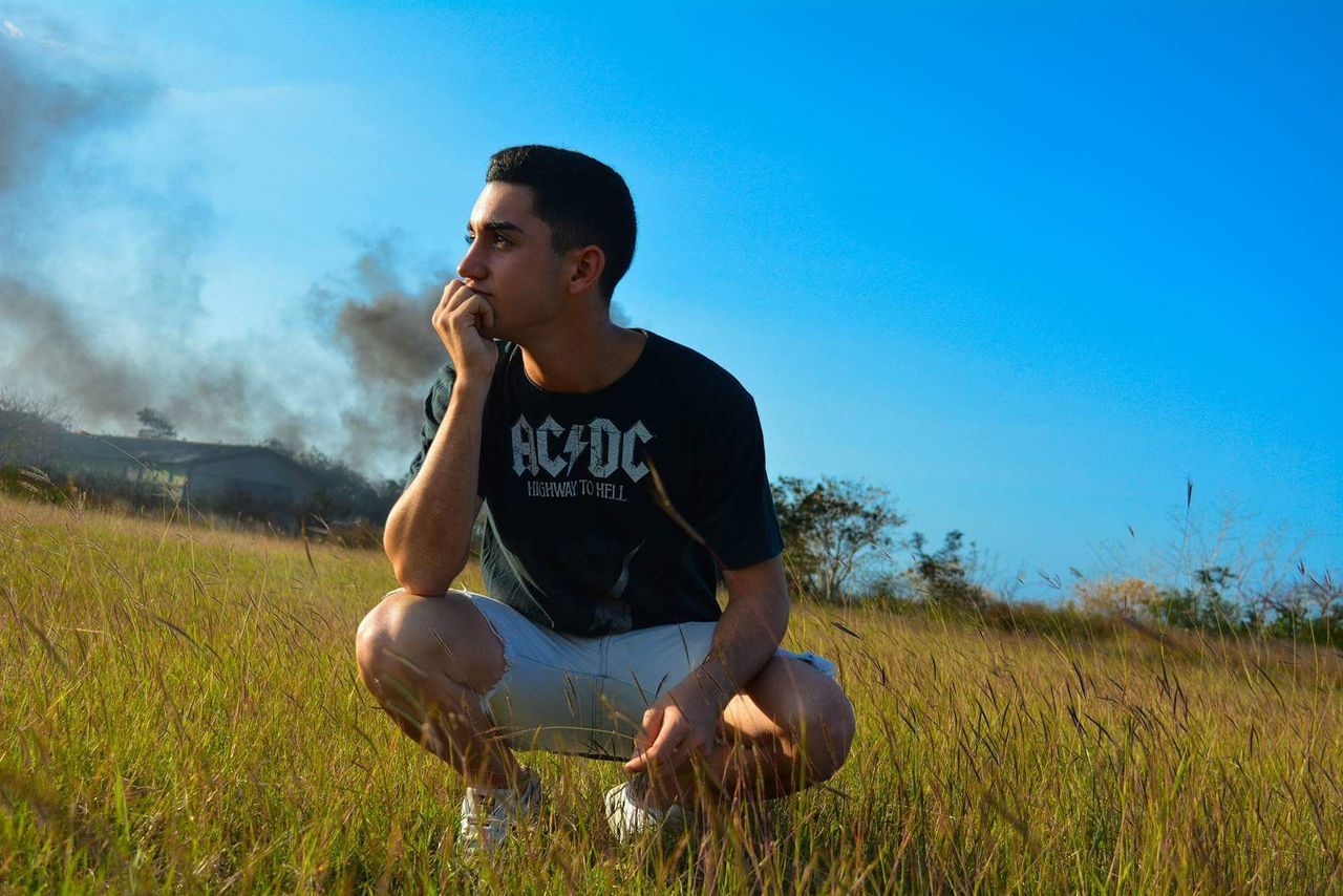 field, one person, casual clothing, land, nature, grass, plant, sky, leisure activity, lifestyles, sitting, real people, day, blue, young adult, front view, looking, looking away, sunlight, contemplation, outdoors, teenager