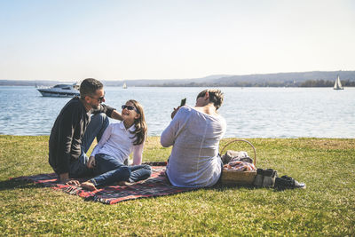 Family enjoying on grassy field by lake against clear sky during sunny day