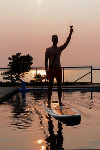 Man holding wineglass while standing on paddleboard in pool during sunset