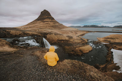 Rear view of man sitting on land by waterfall against sky