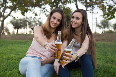 Smiling friends with alcoholic drinks in bottle sitting on grassy field at park