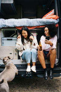 Joyful young multiethnic female friends drinking beer and having fun while sitting on threshold of camper van near dog and enjoying holidays
