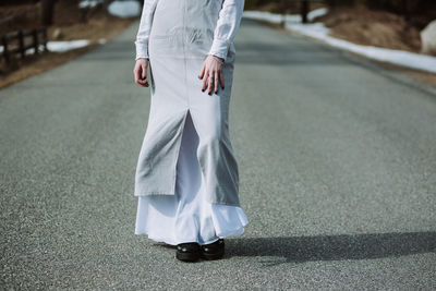 Anonymous young female wearing casual maxi dress strolling along empty asphalt road between evergreen forest in early spring