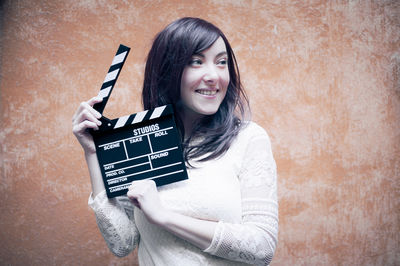 Beautiful woman holding film slate while looking away against orange wall