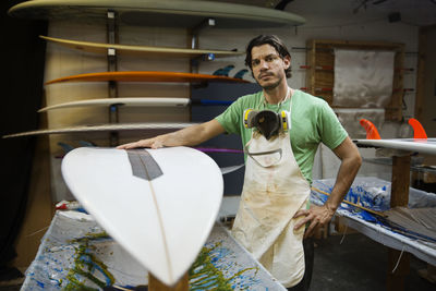 Portrait of serious male worker standing by surfboard in workshop