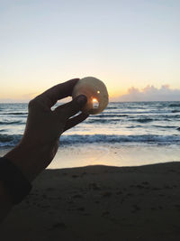 Human hand on sand at beach against sky during sunset