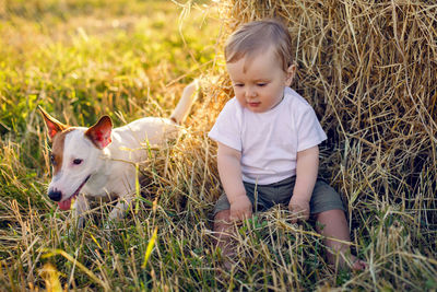 Gay boy kid blonde in white tank top sitting on a field of hay next to  stack with dog during sunset