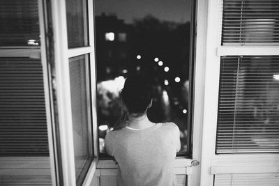 Rear view of thoughtful woman standing by window at night