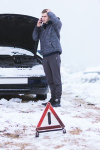 Young man talking over mobile phone while standing by car on snow covered field against sky