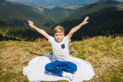 Little boy with arms outstretched sitting in lotus position and enjoying in freedom in nature.