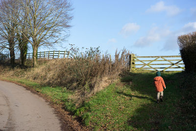 Rear view of a boy in an orange coat walking near a gate on a country road with against a blue sky