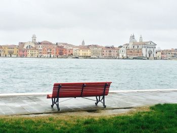 Bench on riverfront with city against cloudy sky